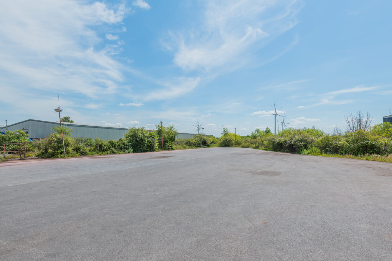Empty paved area with greenery and industrial buildings under a blue sky, wispy clouds, and distant wind turbines.