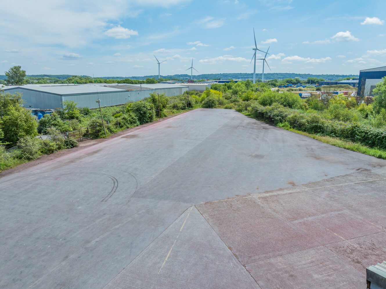Large empty paved car park bordered by greenery, with industrial buildings and wind turbines under a partly cloudy sky in the background.