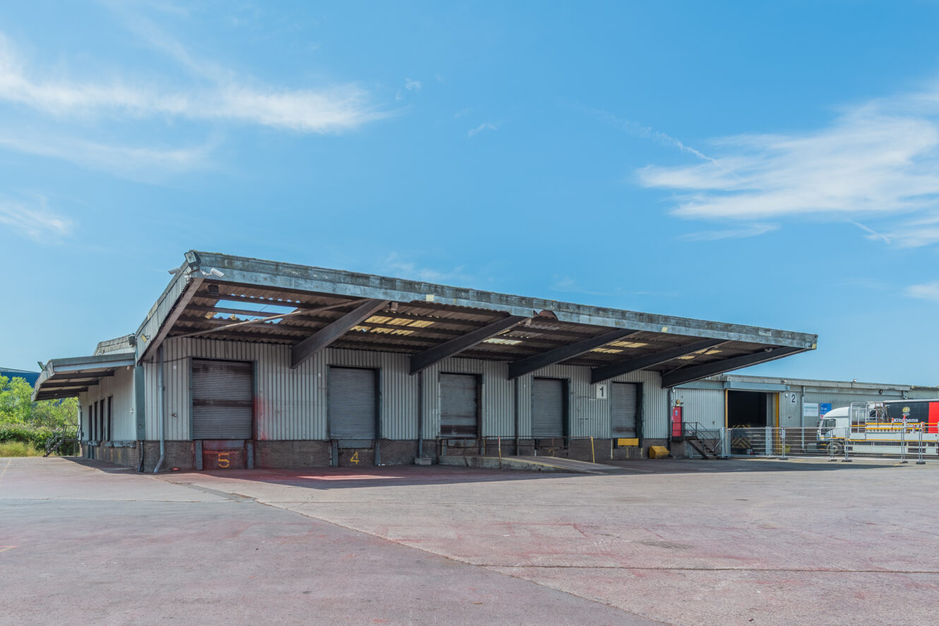 A spacious, empty warehouse loading bay with closed metal doors and a corrugated roof beneath a clear blue sky.