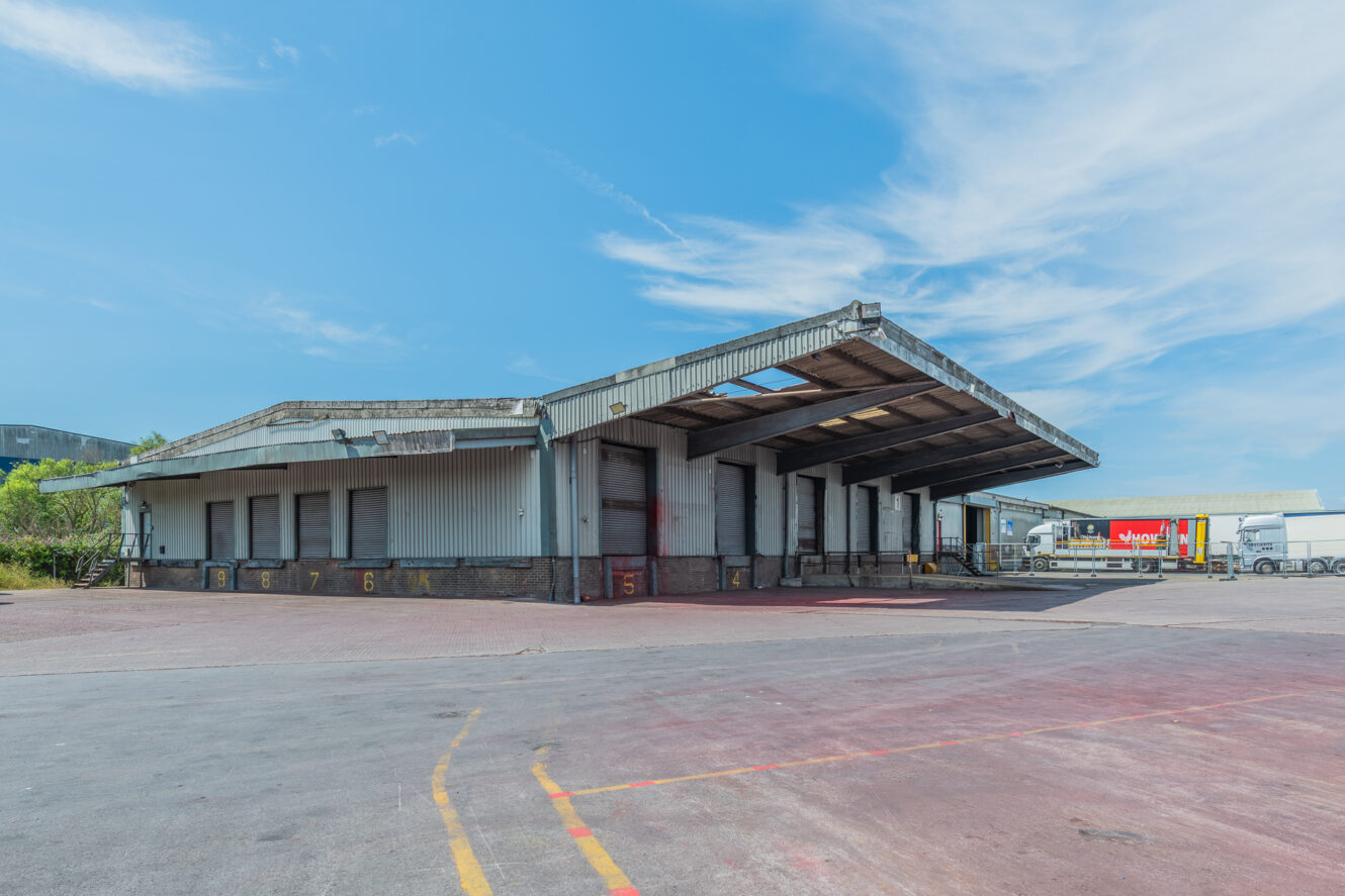 A large, empty industrial loading bay with several bays and a metal roof in an open paved area beneath a blue sky.
