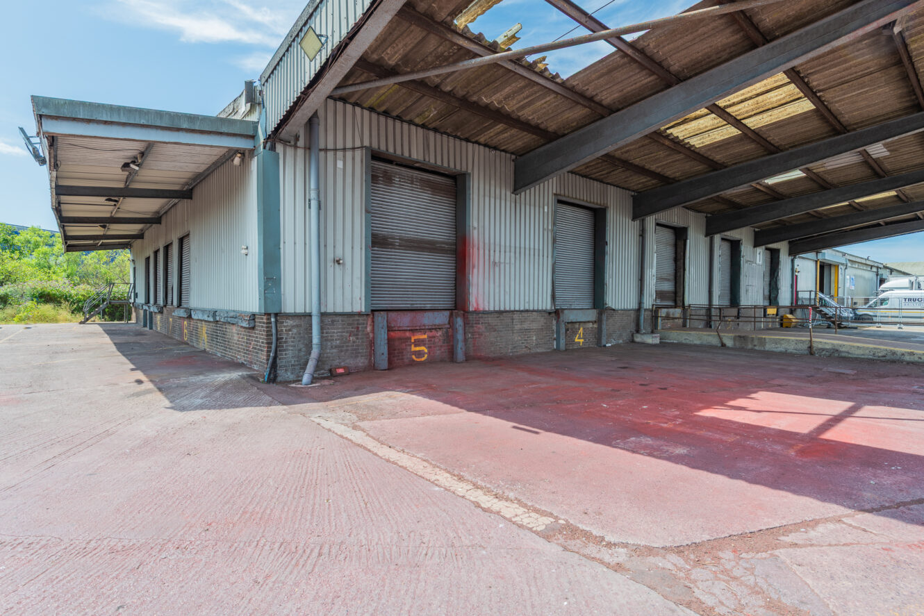 Industrial loading dock with several closed metal roller shutters, concrete bays numbered 4 and 5, and a partially covered roof.