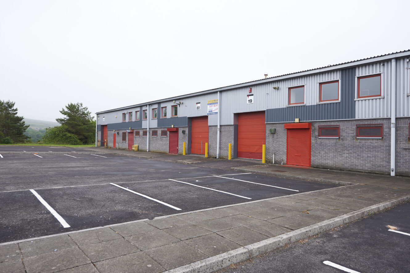 An empty car park in front of a grey industrial building with red doors and windows on an overcast day.