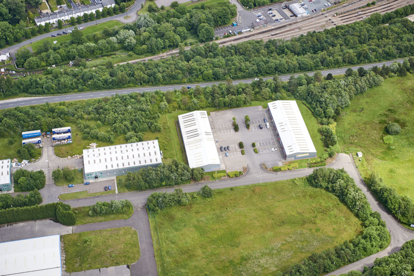Aerial view of industrial buildings with expansive car parks, surrounded by greenery, close to a railway and main road.