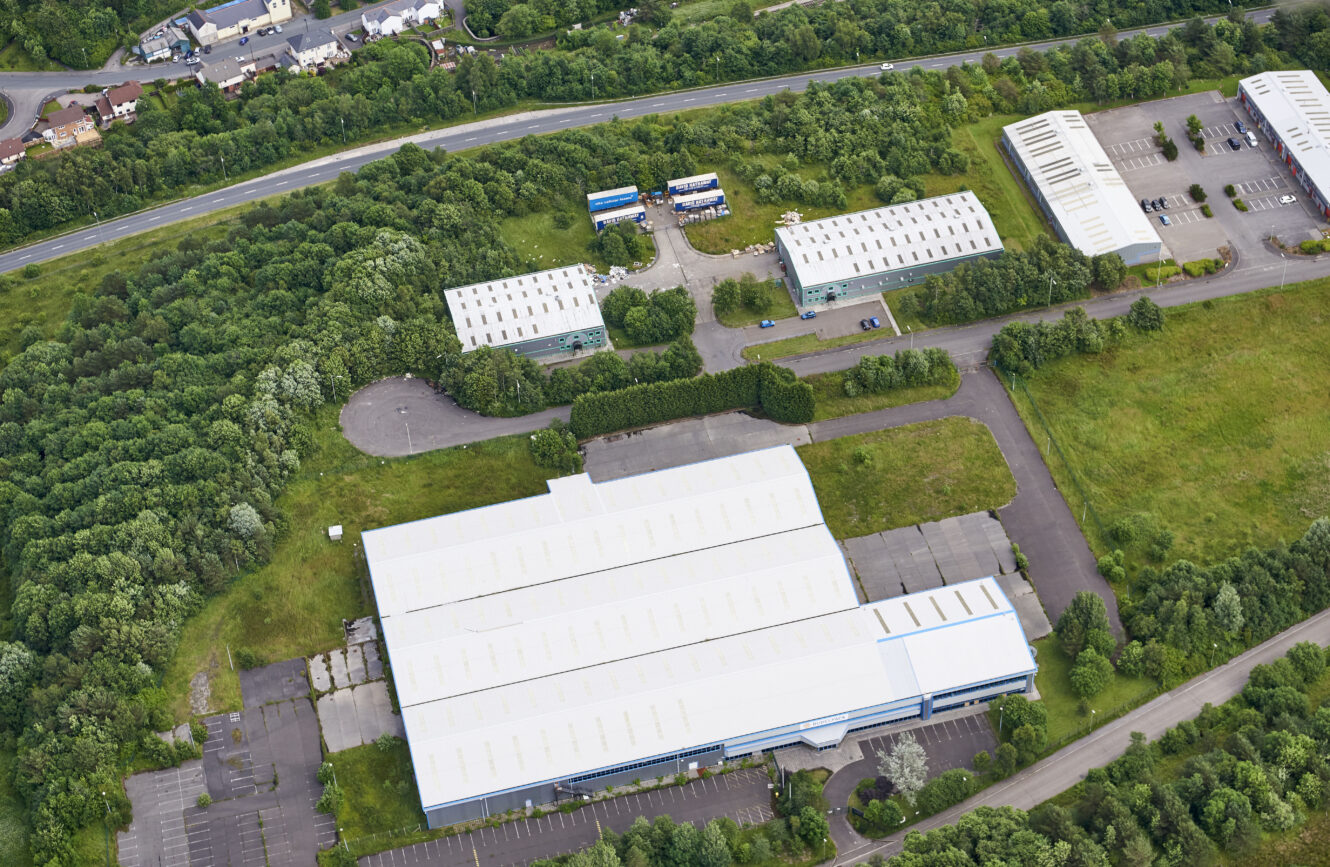 Aerial view of large industrial buildings surrounded by trees, with car parks and a road in the background.