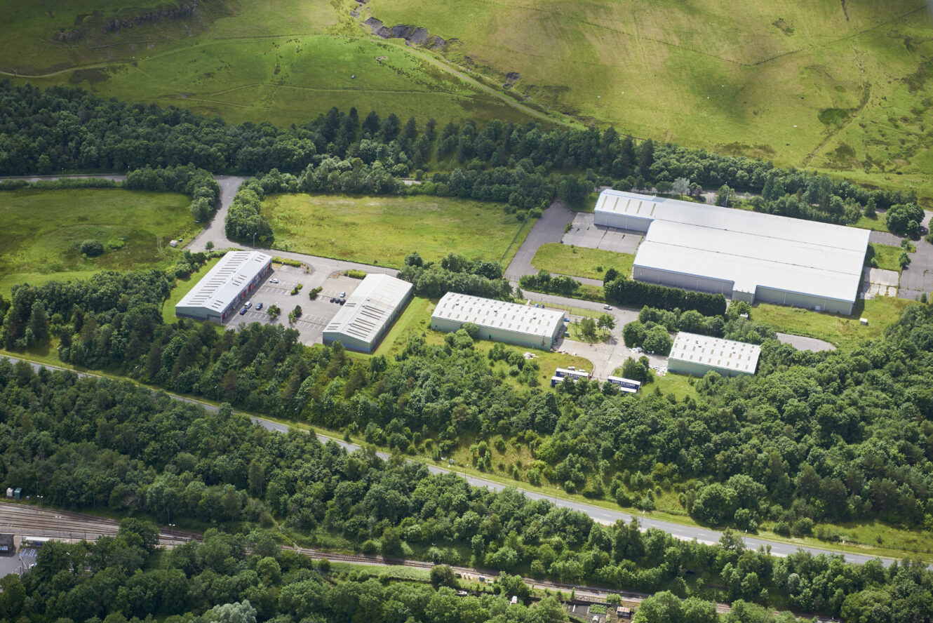 Aerial view of an industrial complex with multiple large warehouses, greenery, trees, and surrounding roads.