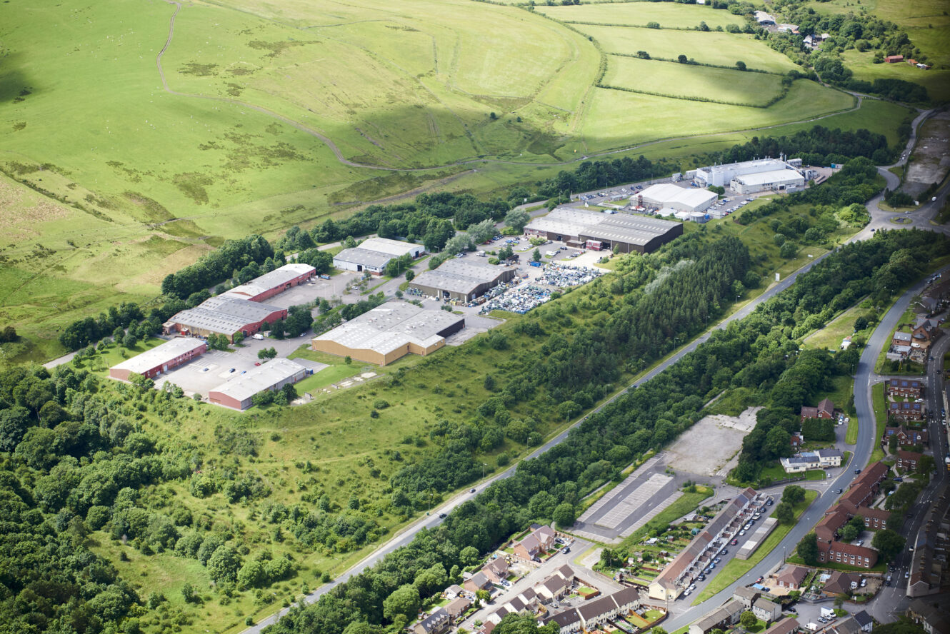 Aerial view of an industrial complex with warehouses, greenery, fields, and nearby residential houses.