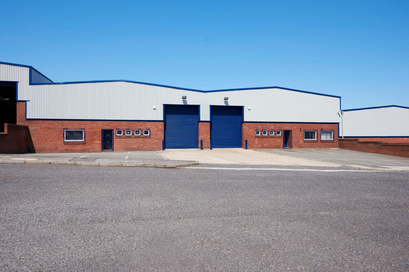 A modern industrial warehouse with two large blue roller shutters, brick lower facade, and corrugated metal upper section under a clear sky.