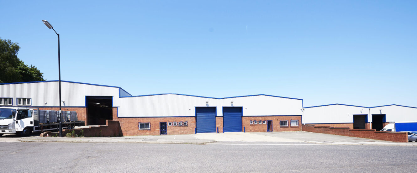 A row of industrial warehouses with brick and white panels, blue doors, and a parked white lorry under a clear blue sky.