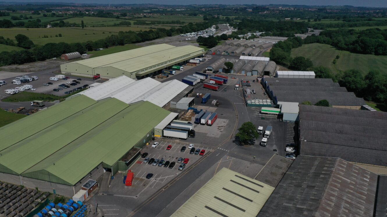 Aerial view of an industrial area featuring warehouses, parked vehicles, stacked shipping containers, and green fields nearby.