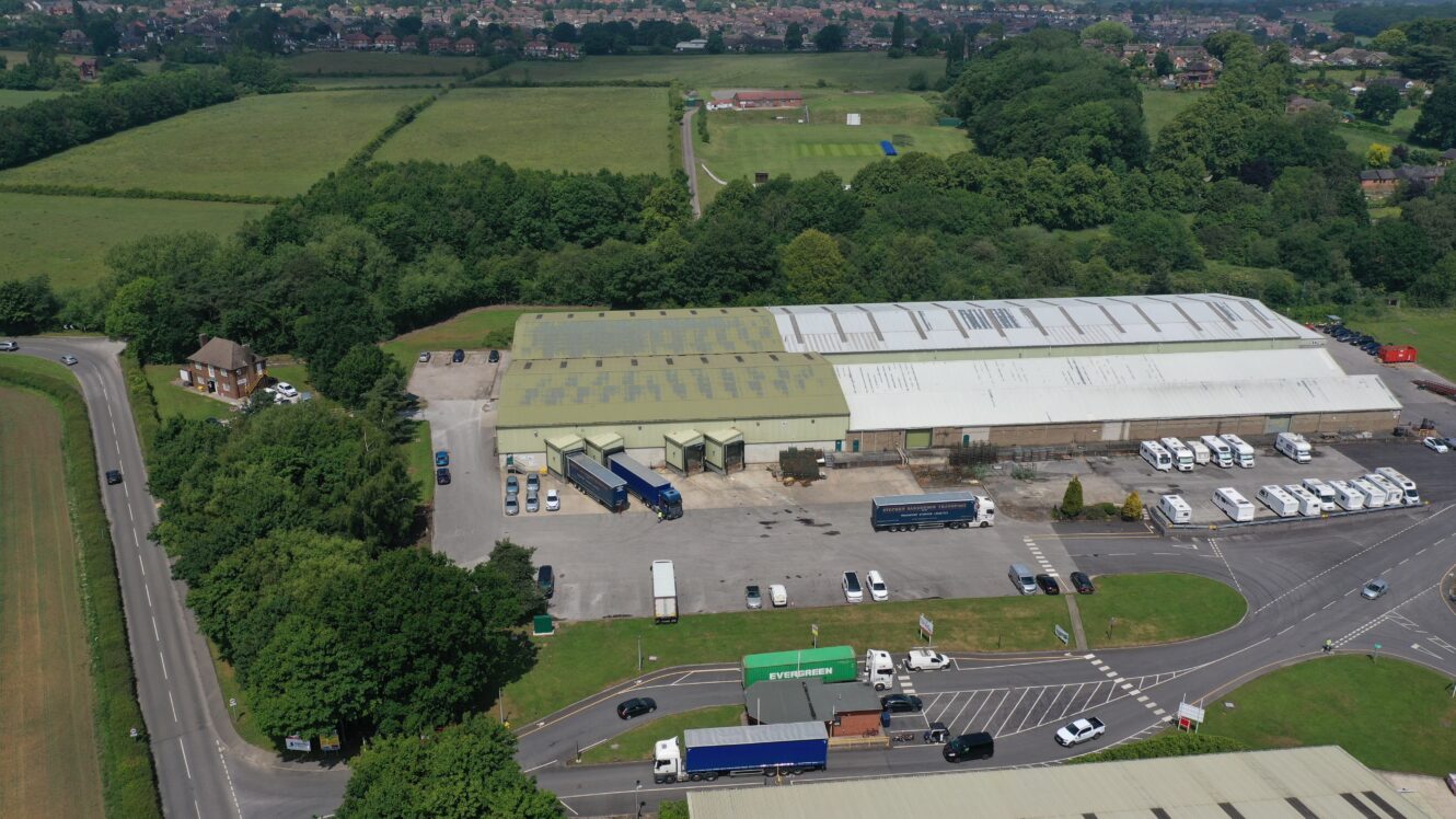 Aerial view of an industrial warehouse complex with lorries and vehicles outside, surrounded by trees, fields, road, and nearby buildings.
