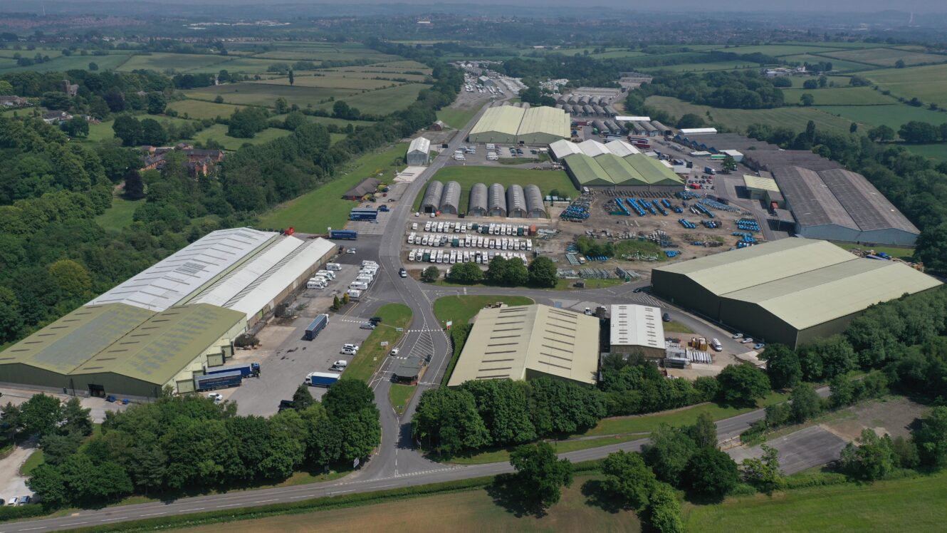 Aerial view of a large industrial estate featuring warehouses, storage units, parked lorries, and nearby greenery in a rural setting.