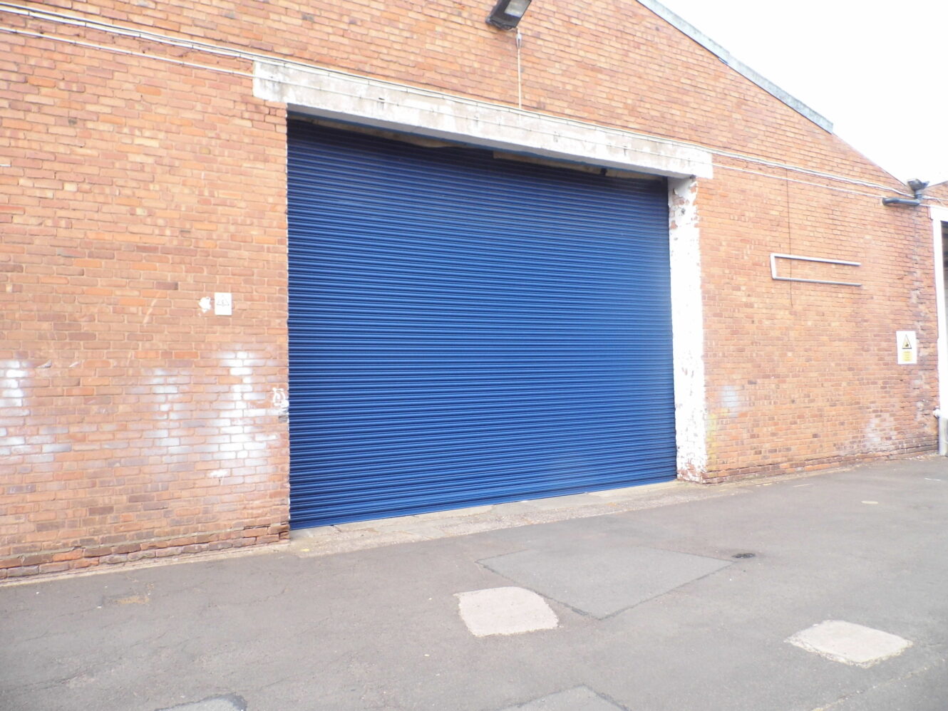 A large blue metal roller shutter door is shut on a brick industrial building, with asphalt paving in front. No people are visible.