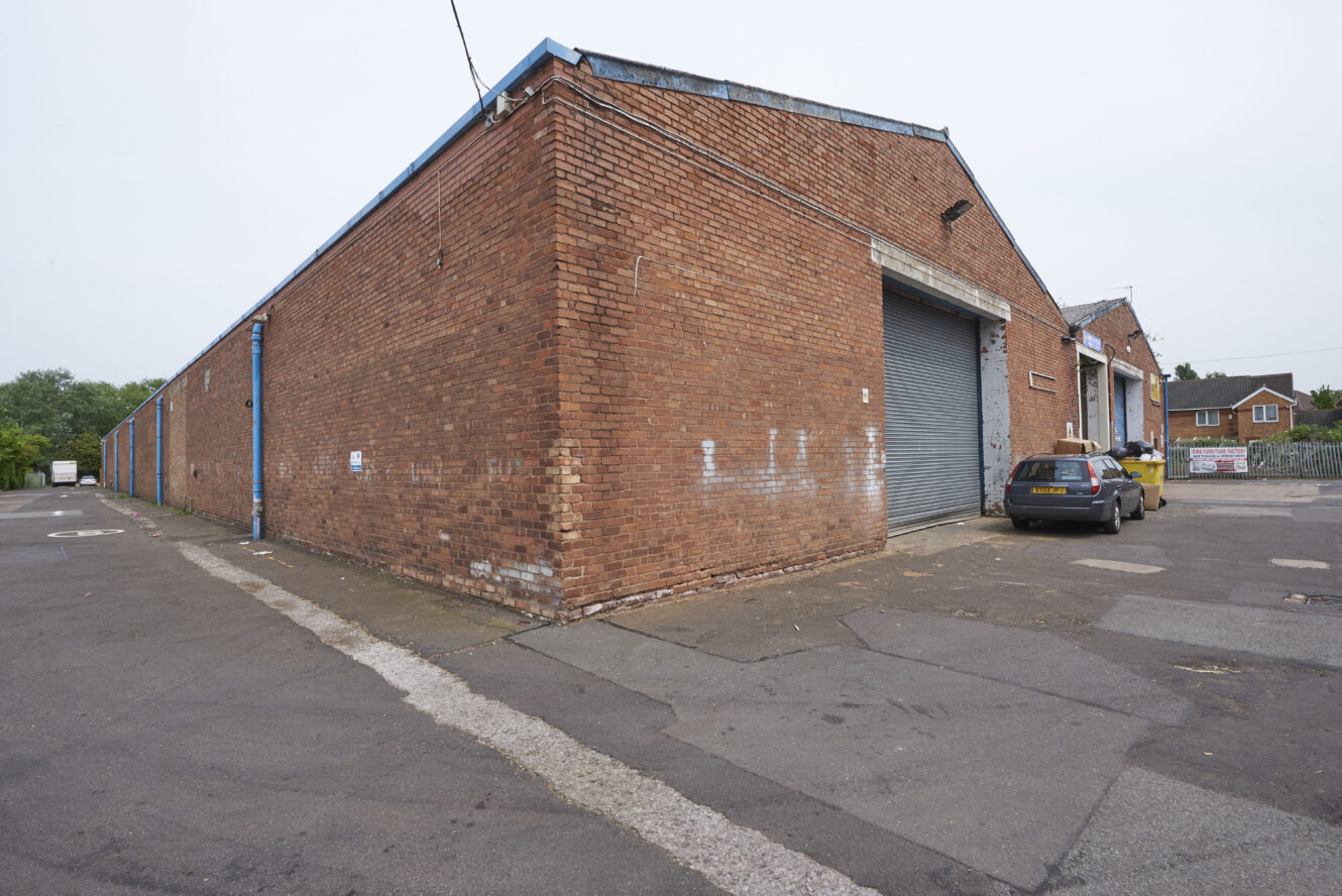 Large brick warehouse with a corrugated metal garage door, parked car, and tarmacked area. Trees and houses are in the background.