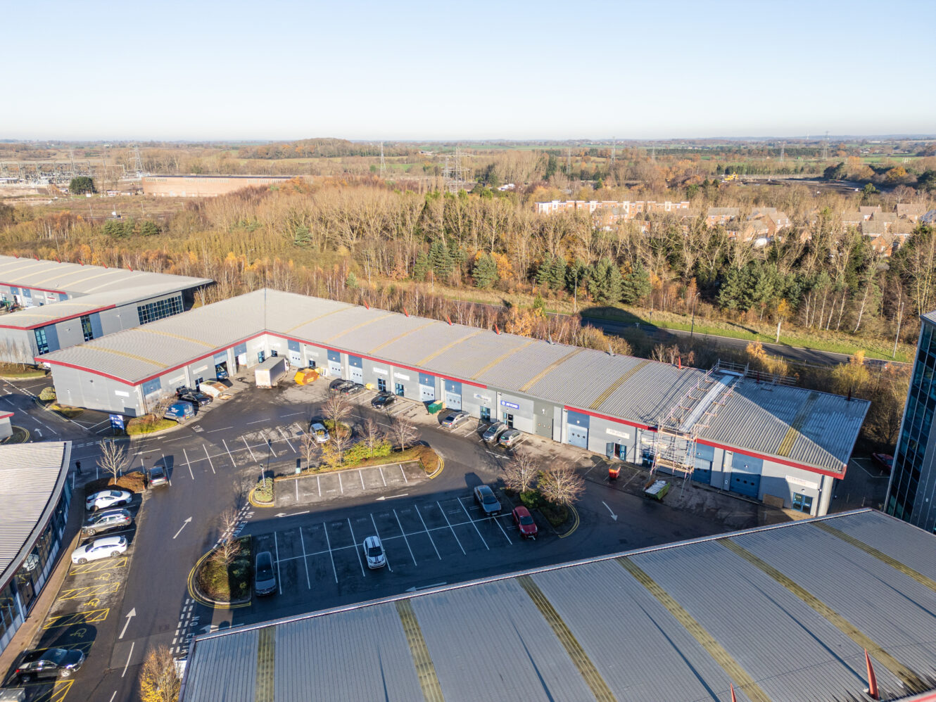 Aerial view of a commercial warehouse complex with parked cars, empty parking spaces, and construction materials; trees and houses behind.