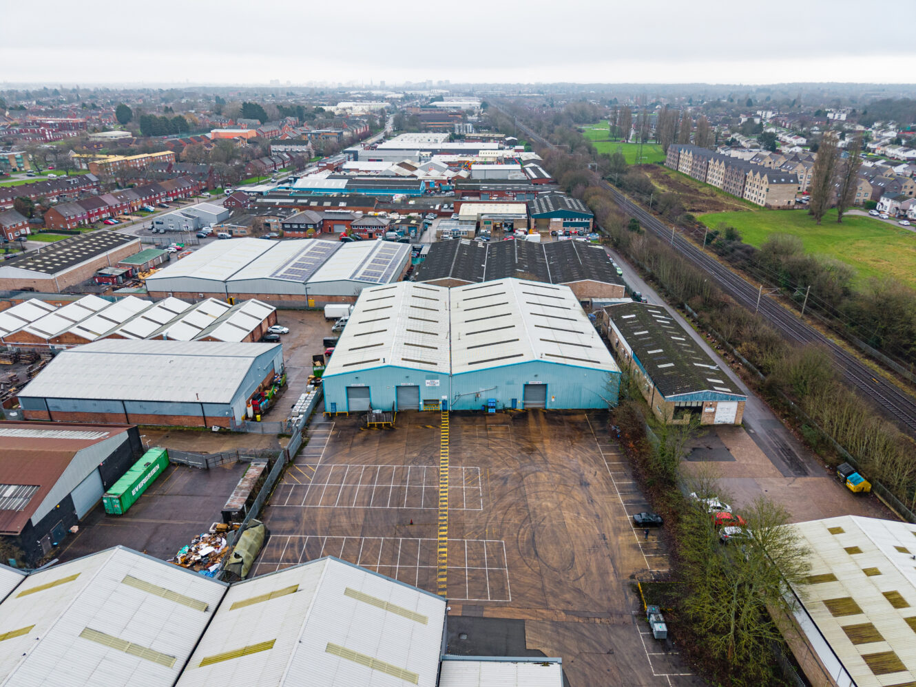 Aerial view of an industrial estate with warehouses, car parks, homes, and railway tracks next to a green area.