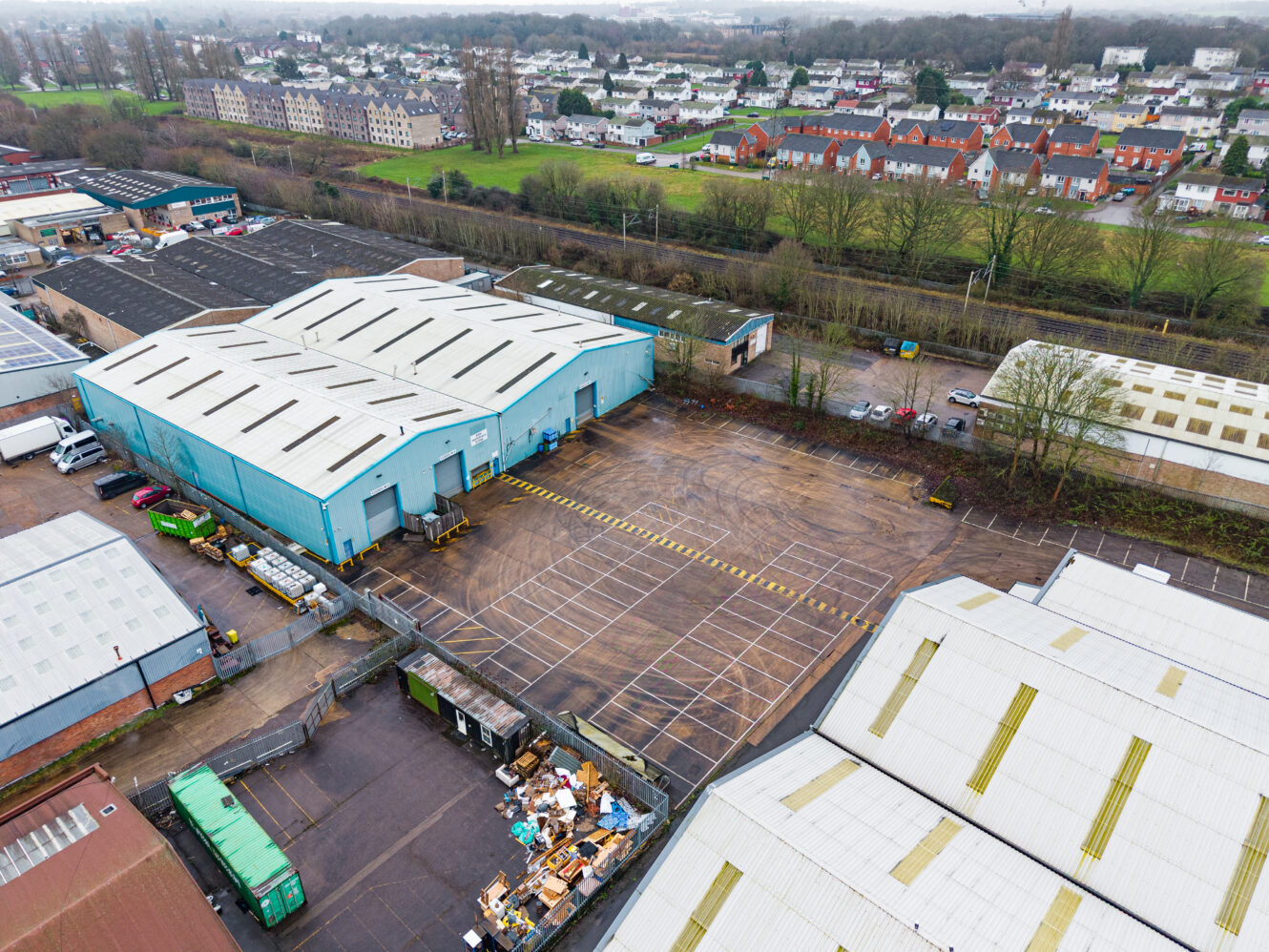 Aerial view of an industrial complex with large warehouses, an empty car park, and nearby residential houses in the background.