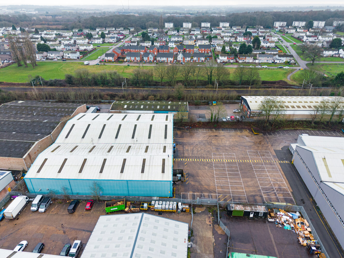 Aerial view of industrial warehouses, parked vehicles, storage areas, and a residential neighbourhood in the distance.