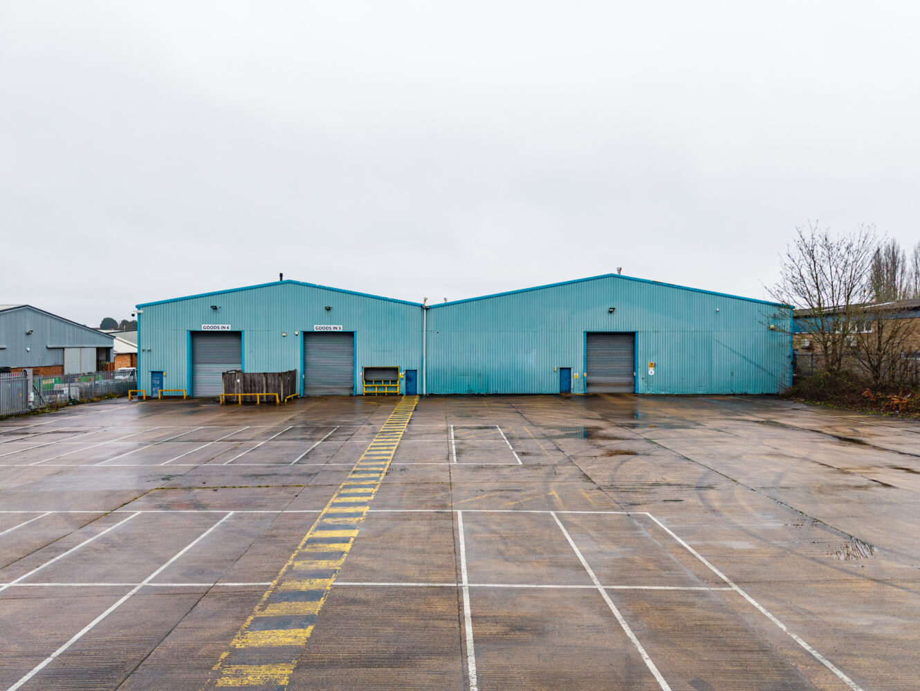 Large blue industrial warehouse with two closed roller shutters, an empty car park in front, and a cloudy sky above.