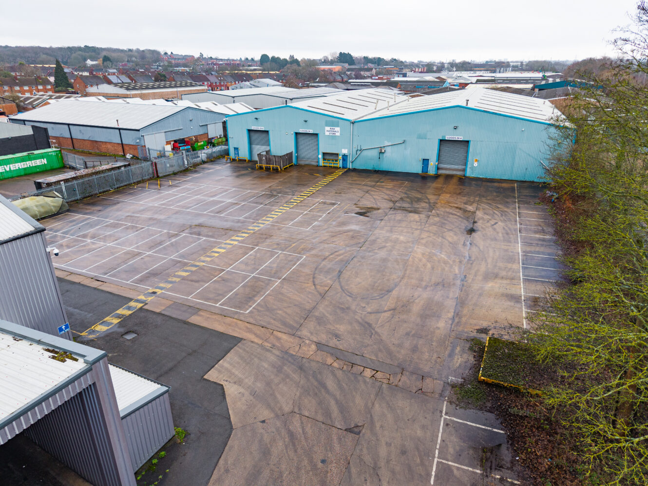 Large industrial warehouse buildings with light blue exteriors and an empty marked concrete car park, photographed on an overcast day.
