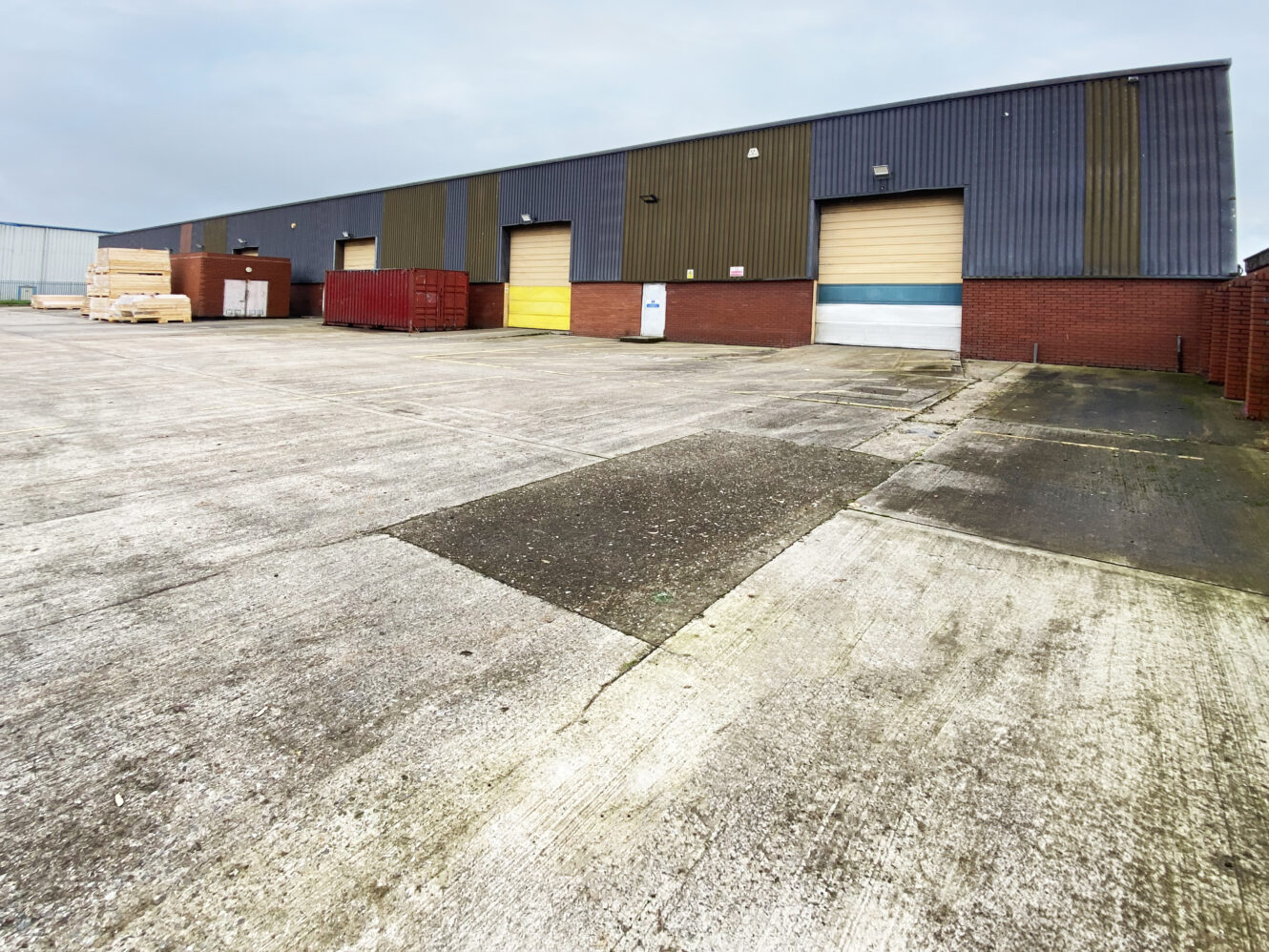 Large industrial warehouse with multiple loading bay doors, red metal containers, wooden pallets, and a wide concrete yard in front.