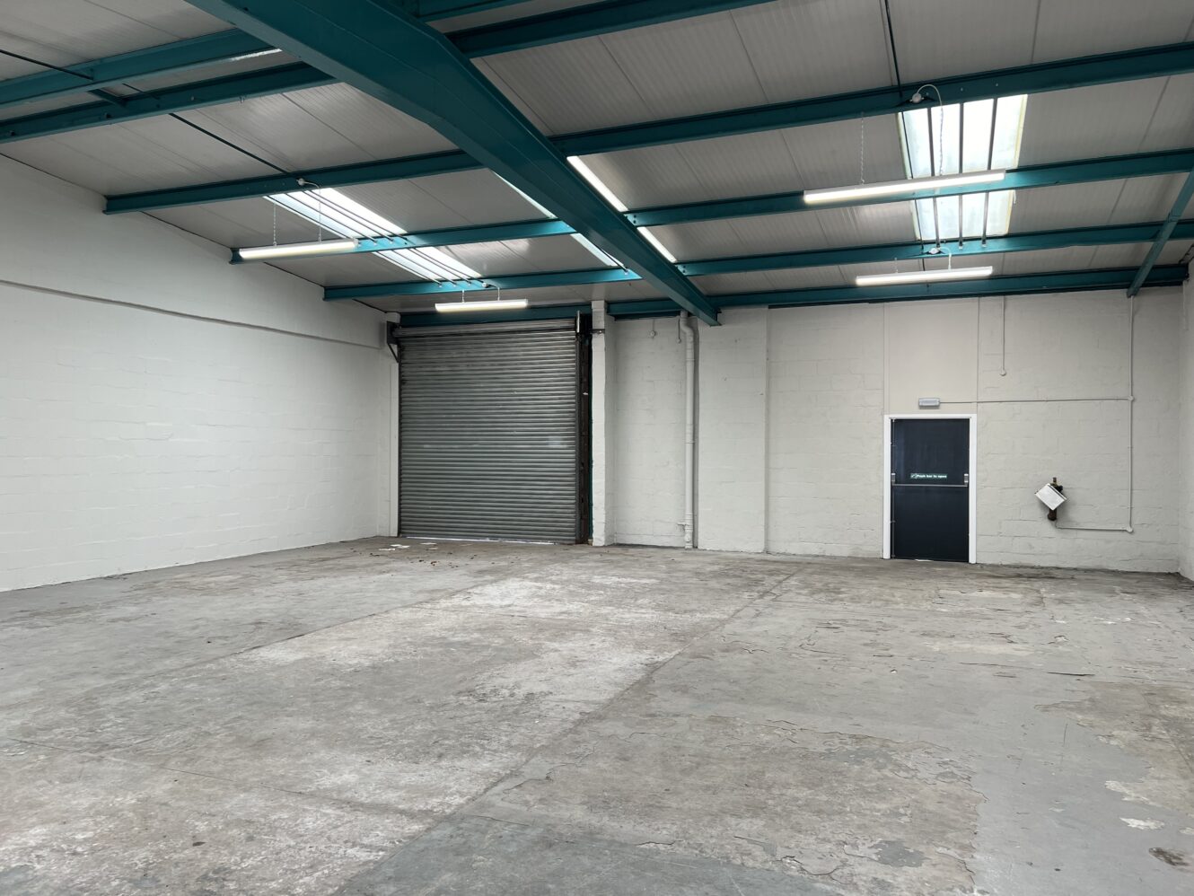 Empty industrial warehouse with concrete floor, white walls, teal ceiling beams, a large roller shutter door, and a single black door on the right. Fluorescent lights hang from the ceiling.