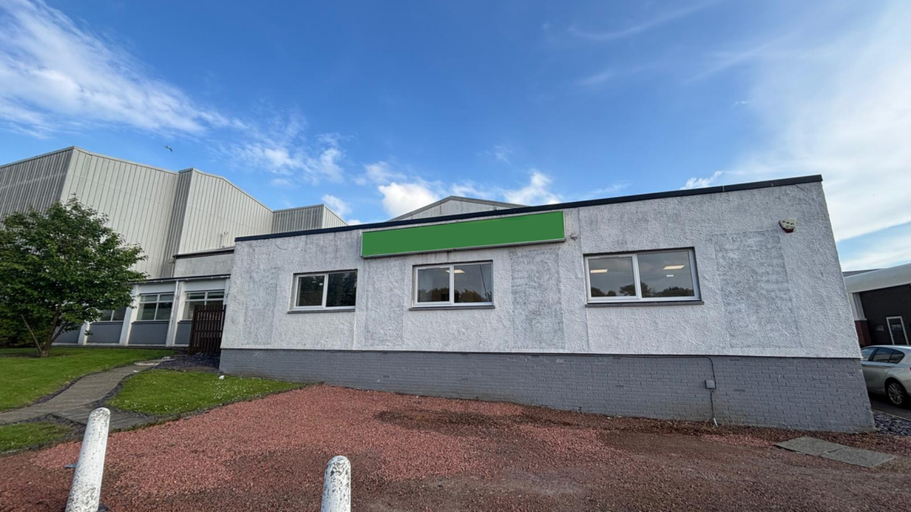 A single-storey commercial building with a green sign, white-grey exterior, and several windows, set against a blue sky with clouds.