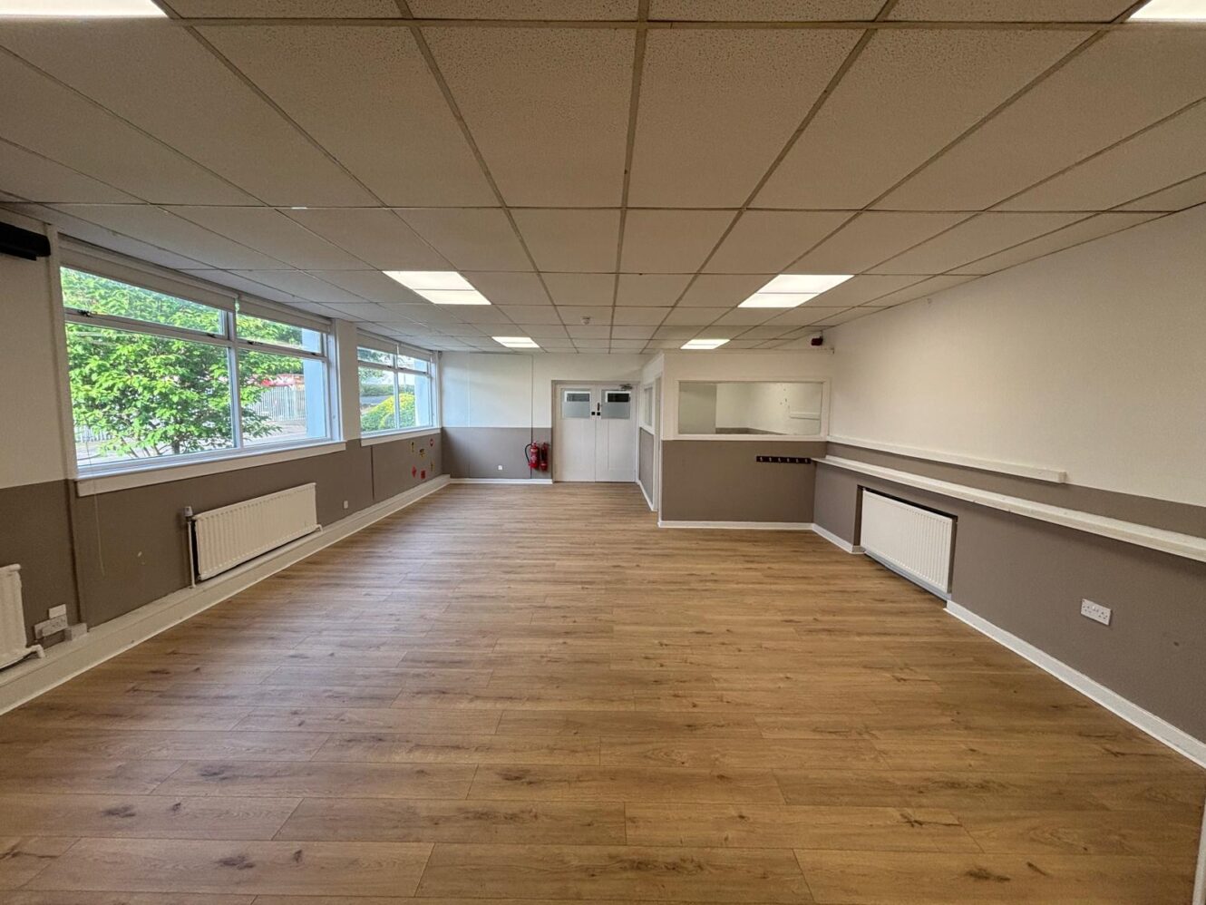 Empty room with wooden flooring, large windows on the left, white ceiling tiles, radiators, and a counter along the right wall. The back wall features a door and a small window.