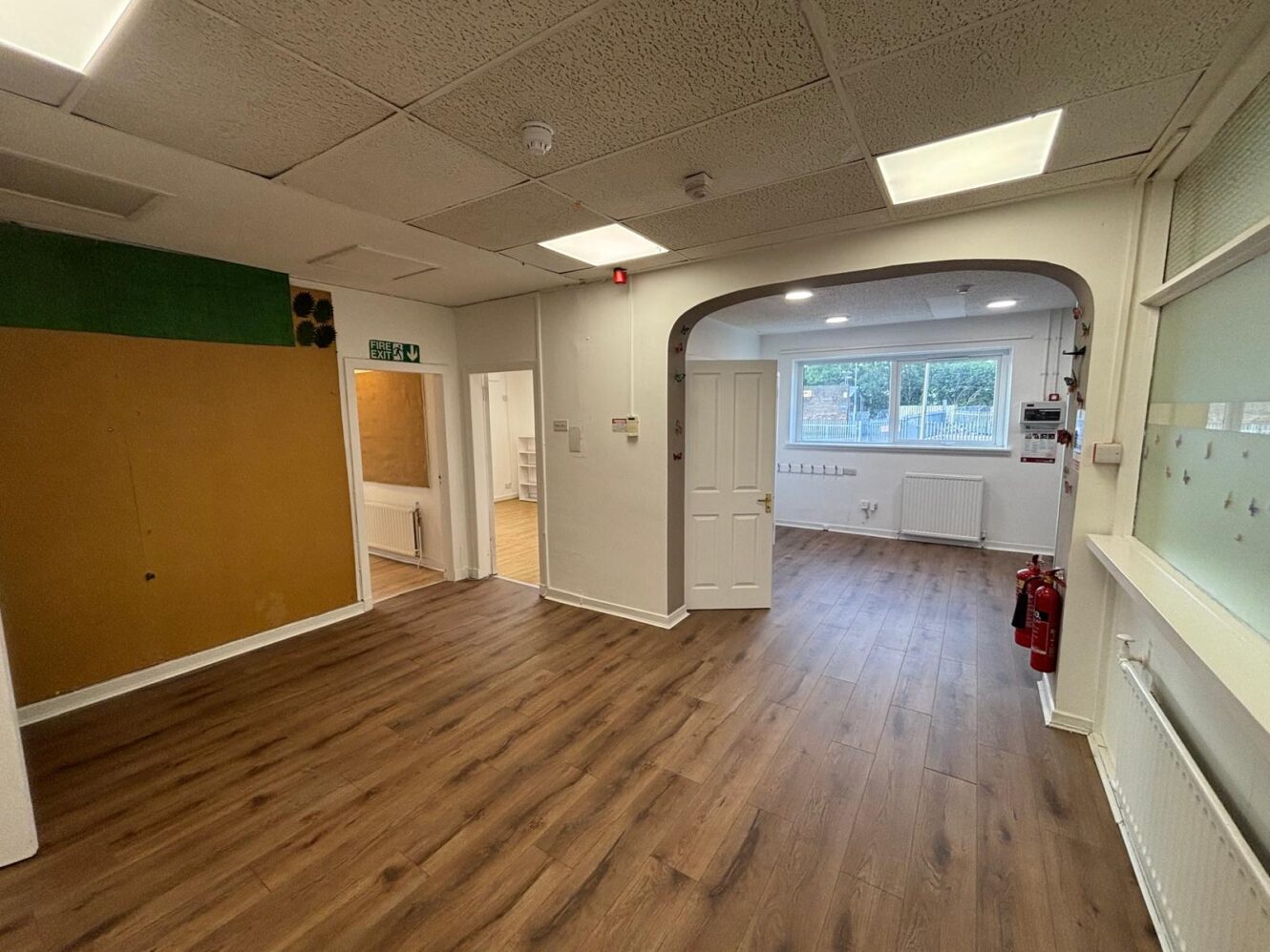 Empty room with wooden flooring, white walls, two fire extinguishers, a large window, fluorescent lighting, and an archway leading to another area.