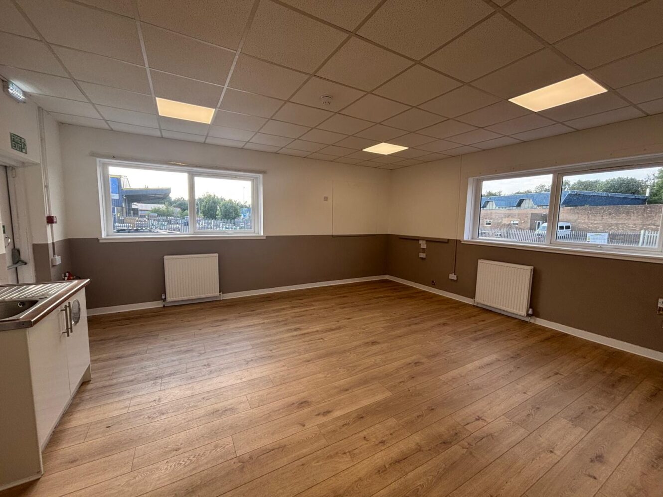 Empty room with wooden flooring, white and beige walls, two large windows, ceiling lights, radiators, and a small kitchen sink area on the left.