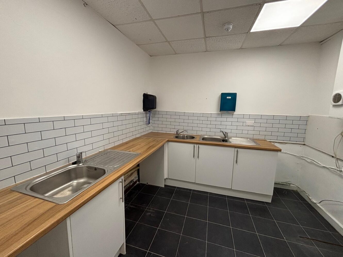 Empty kitchen featuring two sinks, white cupboards, wooden worktops, tiled splashback, black floor tiles, and wall-mounted soap and paper towel dispensers.
