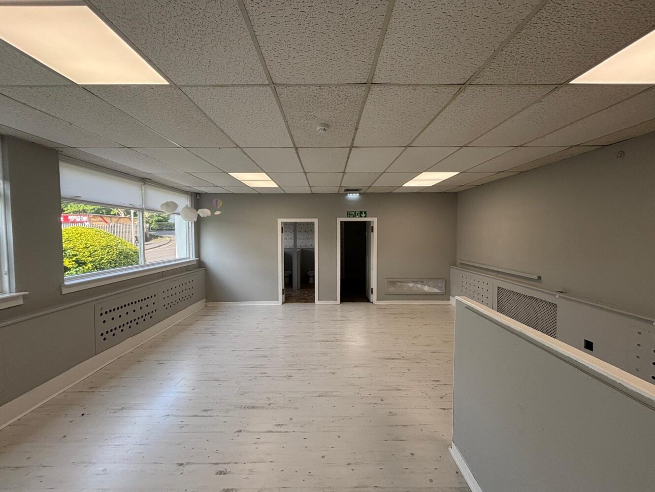 Empty office space with light-coloured wood flooring, grey walls, suspended ceiling with fluorescent lights, large windows on the left, and two doorways at the back wall.