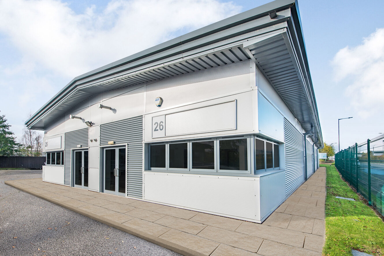 Modern industrial building with a white and grey exterior, featuring the number 26, large windows, and a paved footpath along the side.