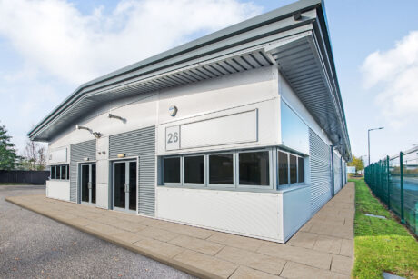 Modern industrial building with a white and grey exterior, featuring the number 26, large windows, and a paved footpath along the side.