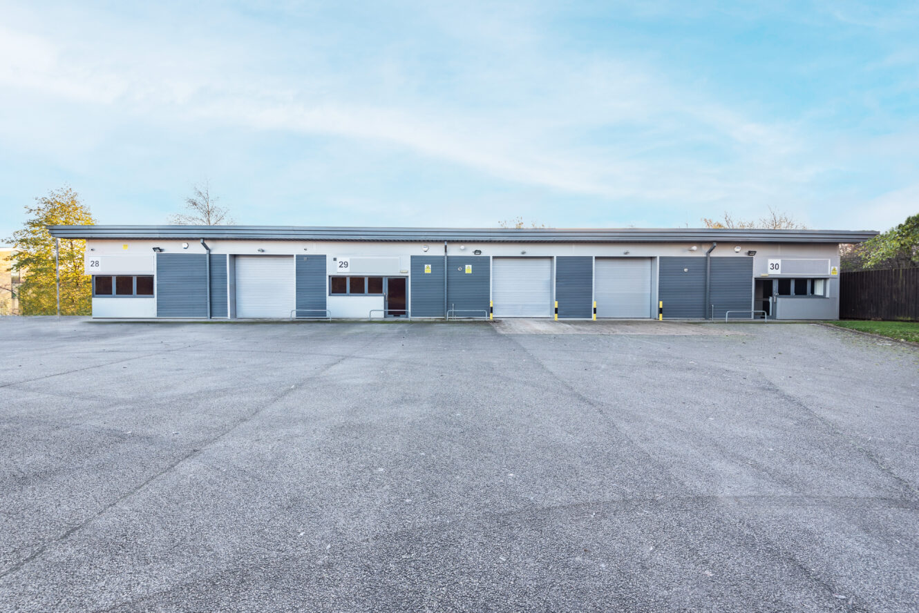 A large, empty tarmac car park in front of a modern industrial building with multiple garage doors and numbered loading bays under a clear sky.