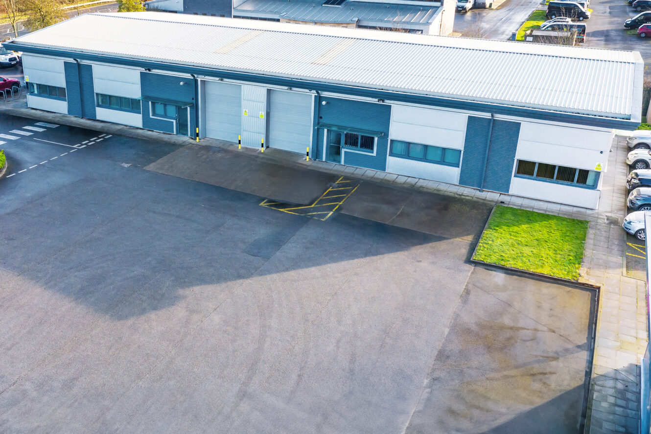 Industrial building with five roller shutters, a central glass entrance, and adjacent parking spaces. A large paved area lies in front, with a small grass verge to the right.