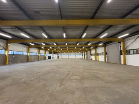 Empty industrial warehouse with yellow steel beams, concrete floor, high ceiling, and exposed fluorescent lighting. Large roller shutter door and windows line the far wall.