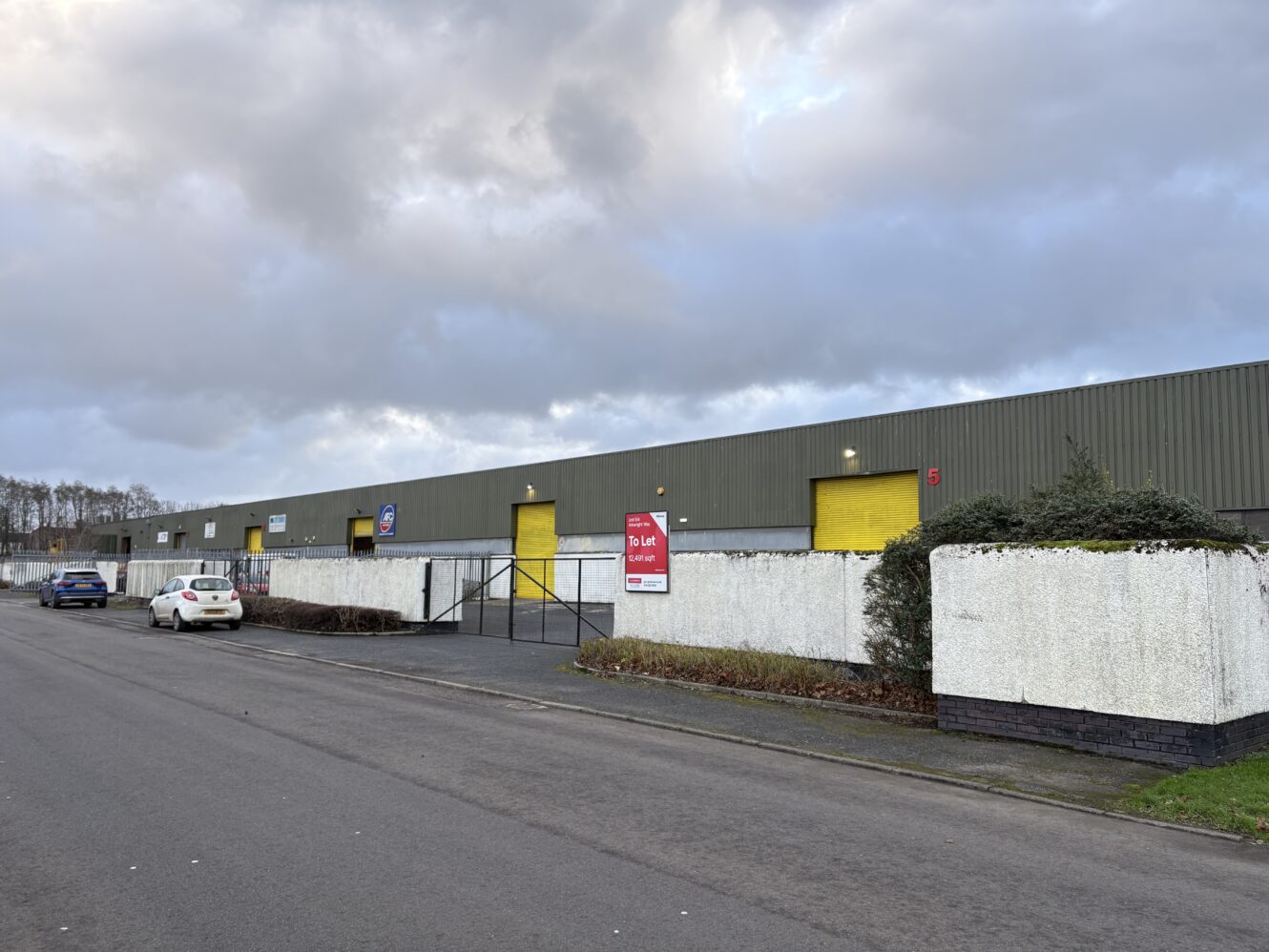 A long, grey industrial building with yellow doors and a To Let sign, bordered by a white wall and bushes, stands next to a street with parked cars under a cloudy sky.