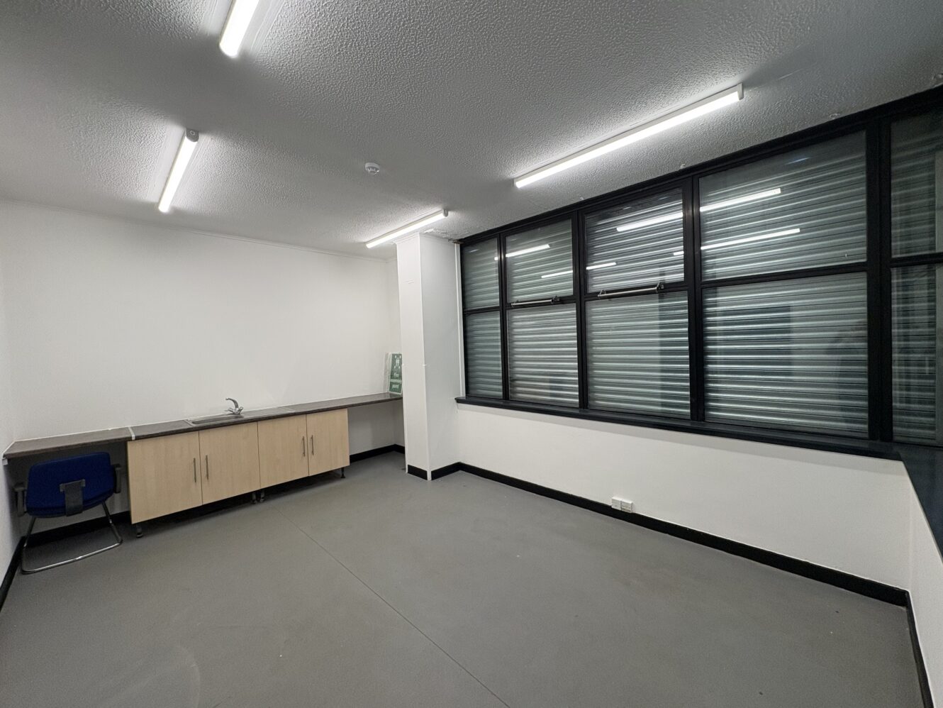 Empty office with grey floor, white walls, a row of cupboards with a small sink, a single blue chair, and large windows covered by horizontal blinds. Bright fluorescent lights on the ceiling.