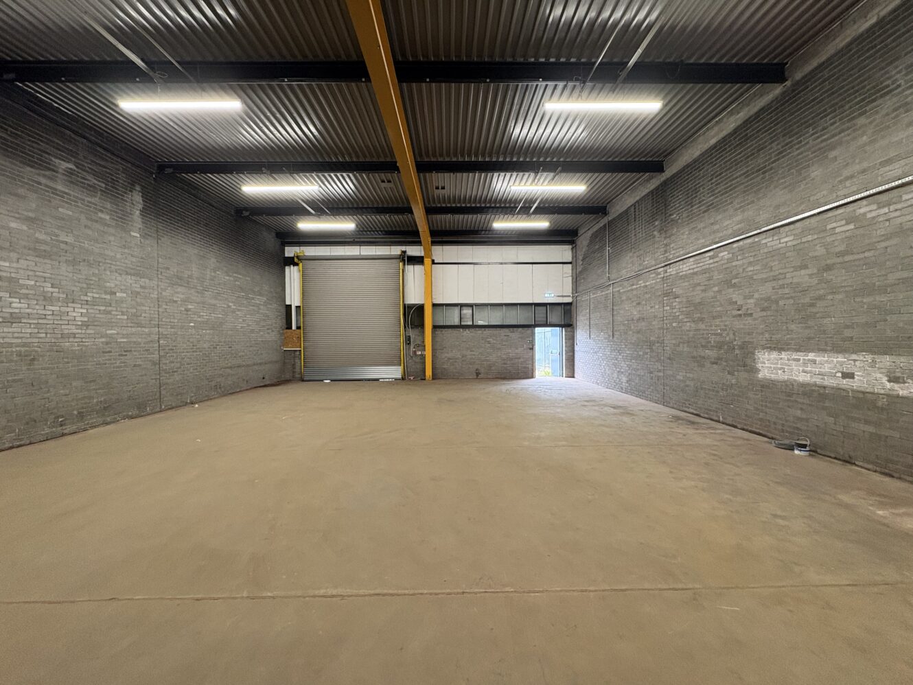 Empty industrial warehouse featuring grey brick walls, a high ceiling, roller shutter door, and overhead yellow crane structure. The concrete floor and fluorescent lighting are also visible.