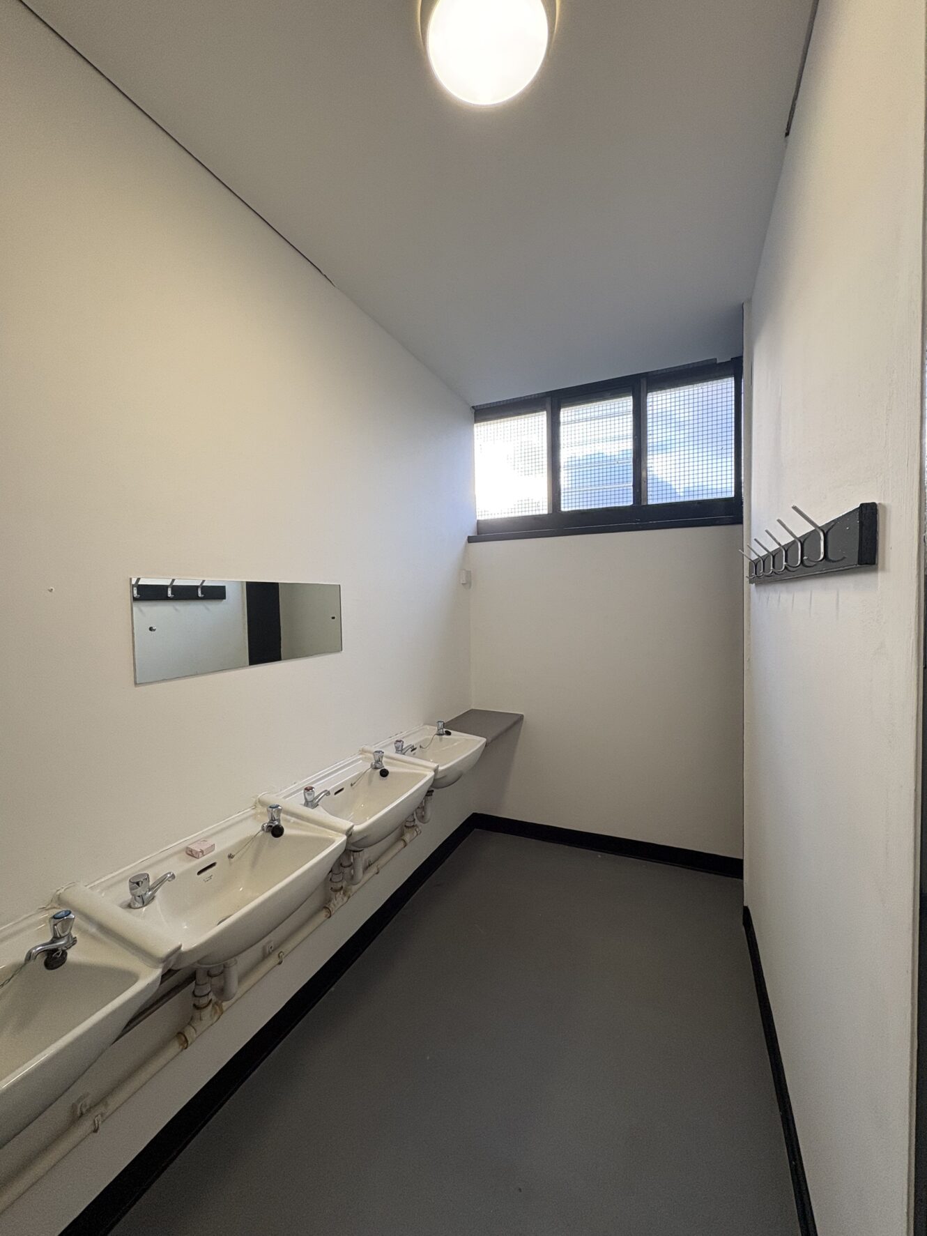 A narrow toilet with three basins, a wall mirror, a coat rack with hooks, and a high window letting in natural light.