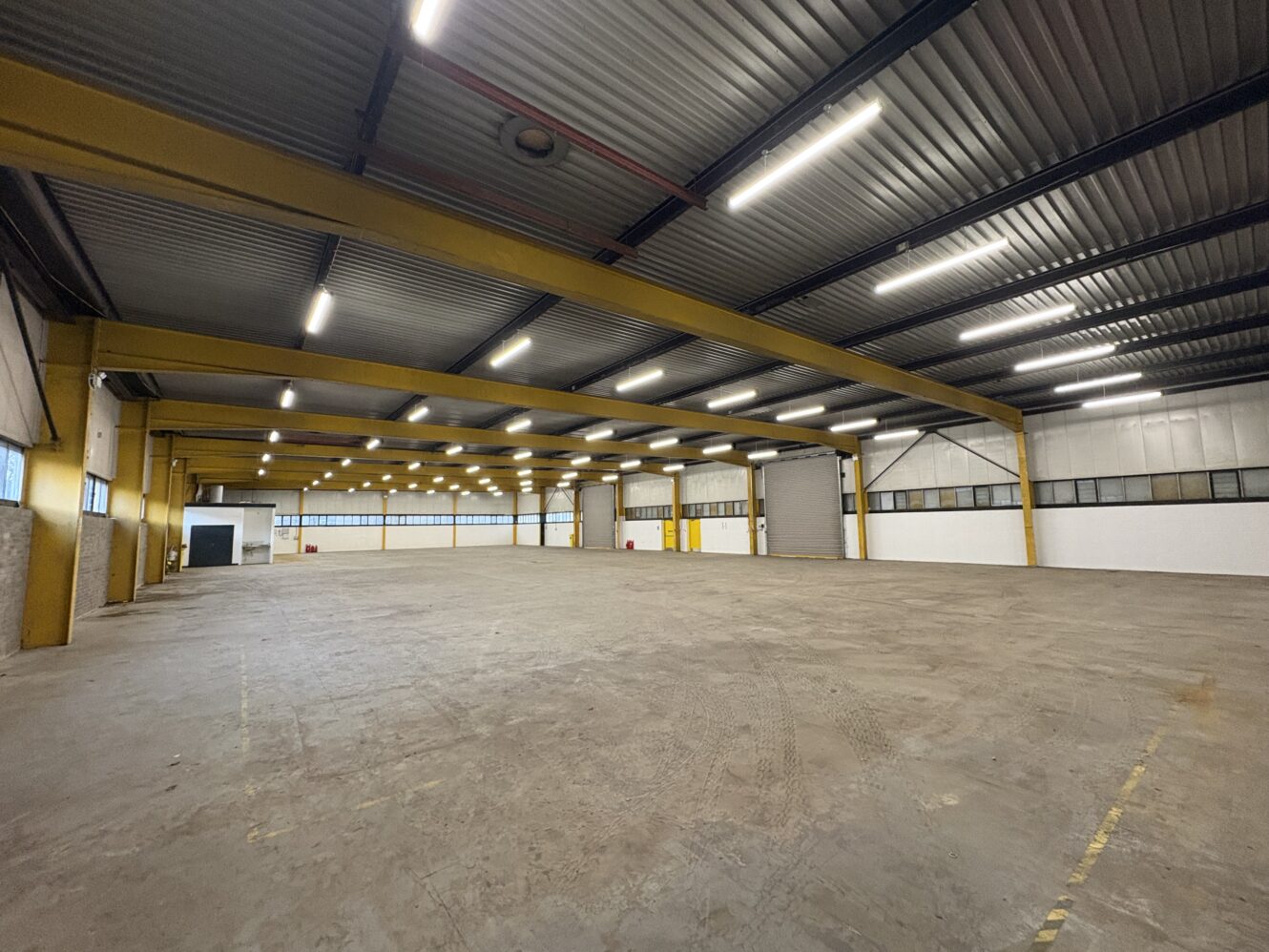 Large, empty industrial warehouse with yellow girders, concrete floors, fluorescent lighting, and multiple windows along the walls.