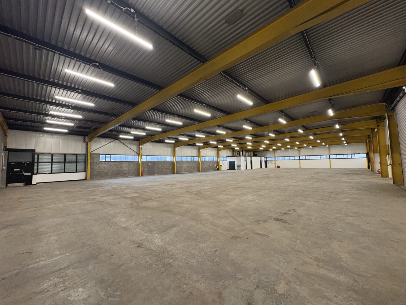 Large, empty warehouse with concrete floor, high ceiling, yellow support columns, fluorescent lighting, and windows along the far wall.