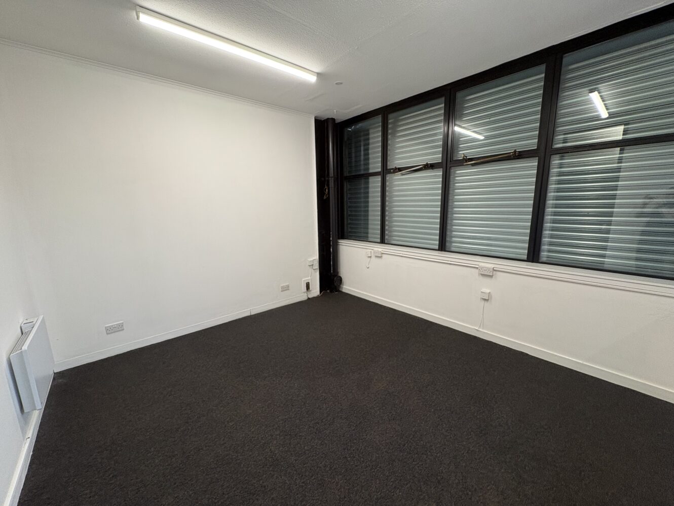 Empty office room with white walls, dark carpet, large windows with blinds, electrical sockets, and overhead fluorescent lighting.