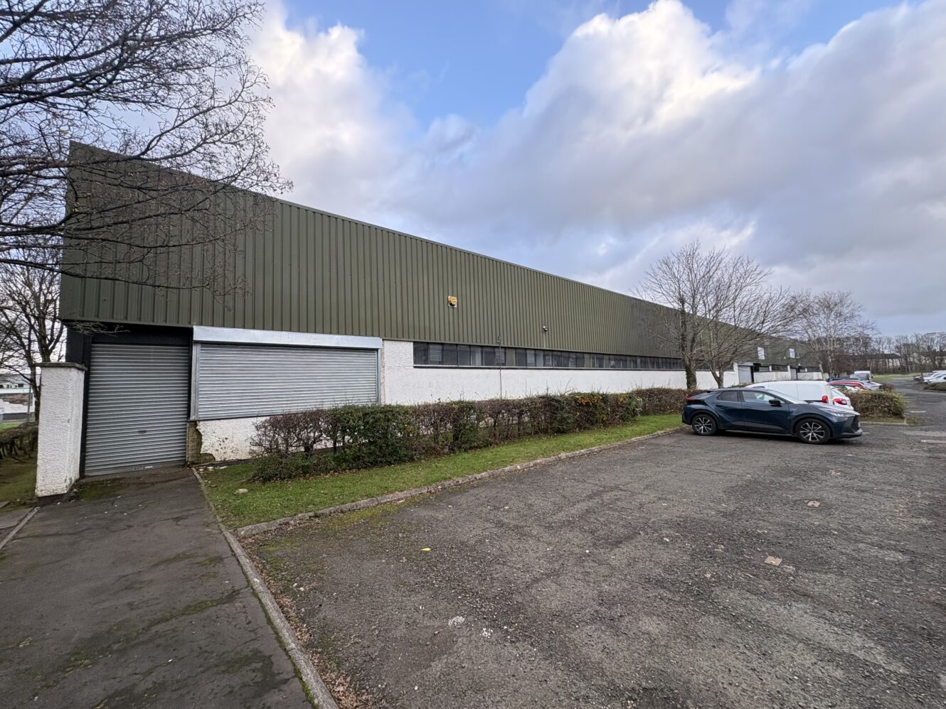 Large industrial warehouse with green metal cladding and white lower walls, next to a mostly empty car park with a few parked cars under a cloudy sky.