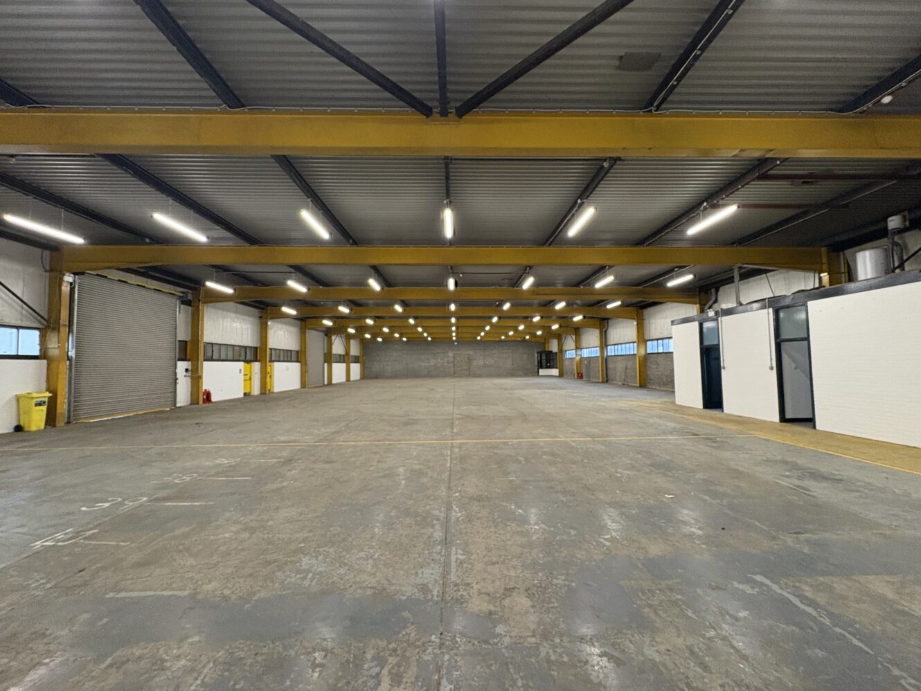 Large empty warehouse with yellow steel beams, concrete floor, fluorescent ceiling lights, and small enclosed office spaces on the right side.