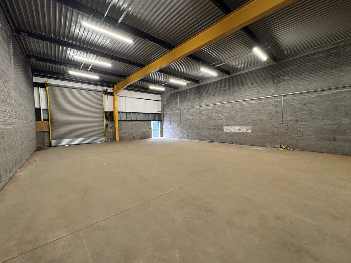 Empty warehouse with a concrete floor, grey brick walls, a large yellow support beam, overhead lighting, and a partially open roller shutter metal door.