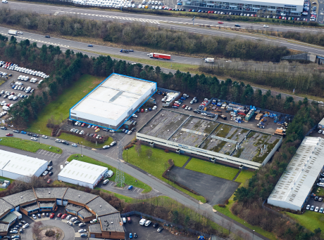 Aerial view of an industrial site featuring warehouses, parked vehicles, a nearby road, and areas of greenery.