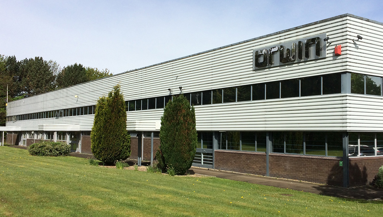 A long, two-storey industrial building with a corrugated metal façade, large windows, and a lawn with trees in front.