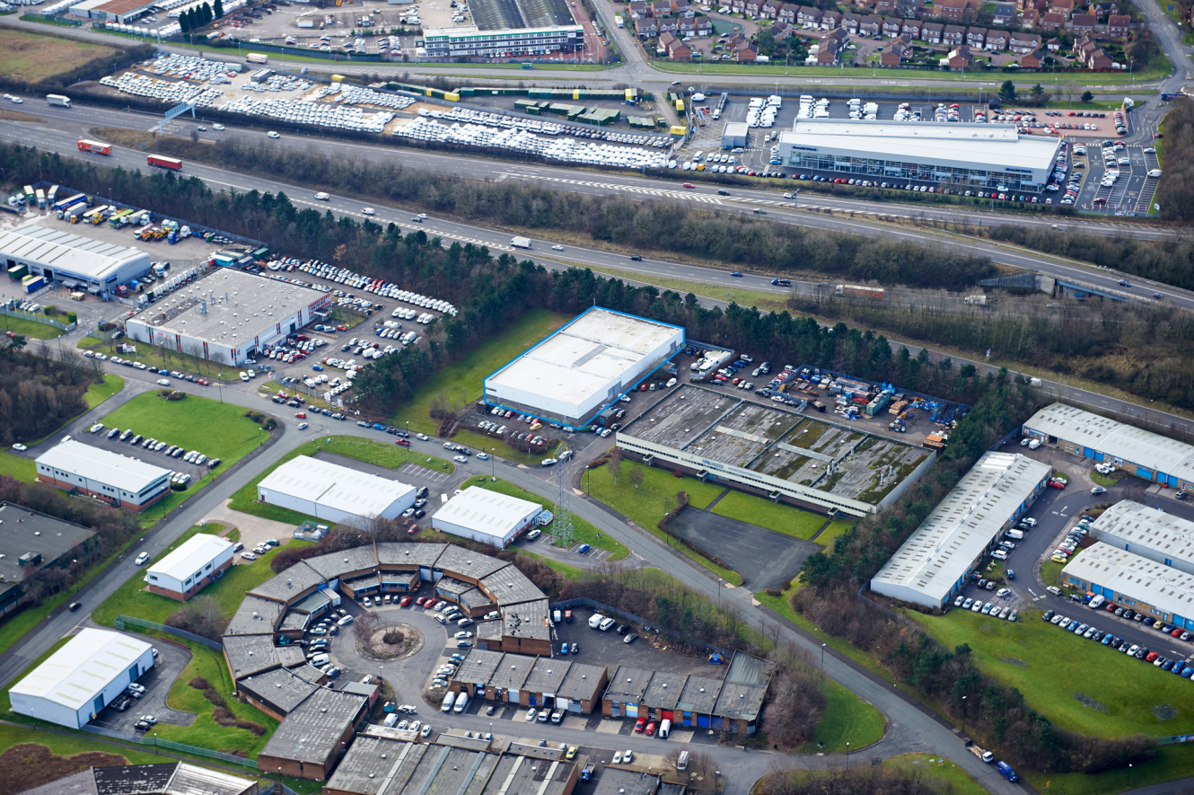 Aerial view of an industrial estate featuring warehouses, circular buildings, car parks, green spaces, and nearby roads and railways.