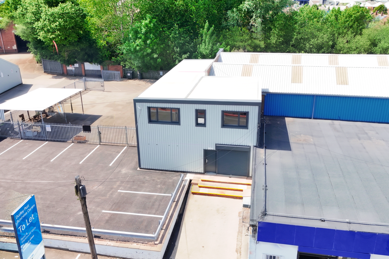 Aerial view of a small industrial building with metal cladding, an adjacent car park, and nearby trees. Yellow ramps lead to a door on the ground floor.
